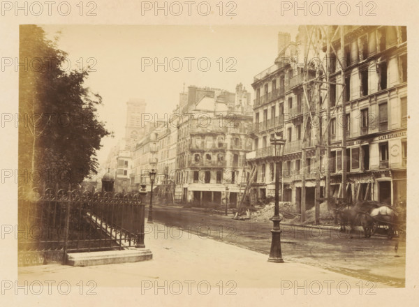 View of destroyed buildings on Rue St Martin, Rue St Martin, photograph, page, Paris, 1871, photographer, paper, albumen print, albumendruk, height 101 mm x width 151 mm, street, theater (building), A sepia-toned street scene showing a row of multi-story buildings with visible damage and scaffolding against a pale sky, ornate wrought-iron balconies and shuttered windows on the facades, piles of debris and wooden supports at the bases of some buildings, a tall ornate street lamp in the foreground and another further down the street, a decorative iron fence and leafy trees along the left side, a domed kiosk set back near the buildings, and a horse-drawn cart partially visible at the lower right.