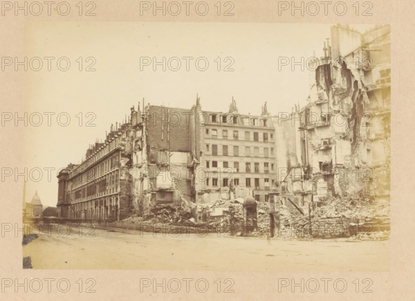View of destroyed buildings on the Rue du Faubourg Saint-Honoré and the Rue Royale, Fabourg St Honoré & Rue Royale, photograph, page, Rue Royale (Paris), 1871, photographer, Paris, paper, albumen print, height 101 mm x width 151 mm, street, theater (building), A wide sepia-toned street scene showing a row of tall multi-story buildings, with several façades partially demolished exposing interior floors and rubble piles in front, a more intact building centered with rooftop dormer windows and shuttered windows, architectural details like chimneys and vertical pipes along the rooftops, a damaged stone wall and scattered debris filling the foreground, a small enclosed structure near the rubble at street level, and a dome-shaped building visible in the distant left background.
