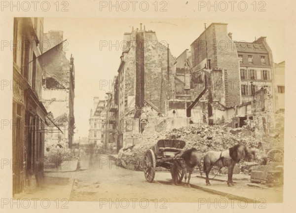 Rue du Bac & Rue de Lille, View of a horse cart in front of destroyed buildings the Rue du Bac and the Rue de Lille, photograph, page, 1871, photographer, paper, albumen print, albumendruk, height 101 mm x width 151 mm, street, two-wheeled, animal-drawn vehicle, theater (building), Paris, A narrow urban street flanked by tall, weathered buildings with exposed brick and plaster, many showing damaged or partially demolished facades, rubble piled along the right side of the street, a wooden cart with large spoked wheels hitched to two horses standing near the rubble, a few indistinct figures further down the street, awnings and shopfronts visible on the left, remnants of interior walls and staircases visible in the ruined structures, and a distant multi-story building seen at the end of the street.