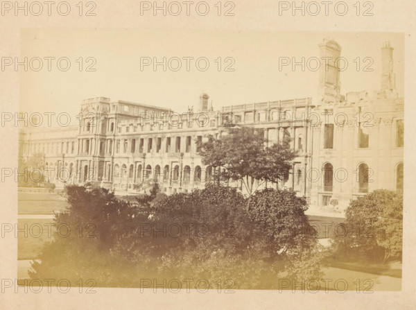 View of the Tuileries Palace in Paris, Palais des Tuileries - vue extérieure, photograph, page, Tuileries Palace, 1871, photographer, Paris, paper, albumen print, height 101 mm x width 149 mm, front facade, palace, A wide sepia-toned photograph of a large classical building with rows of tall arched windows and ornate columns, showing a long facade with multiple stories and decorative stonework, a partially ruined tower rising above the right side, and trees and shrubbery in the foreground partially obscuring the lower part of the building.