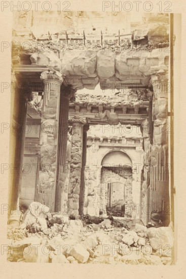 View of the ruined interior of the Tuileries Palace in Paris, Palais des Tuileries - vue interiore, photograph, page, Tuileries Palace, 1871, photographer, Paris, paper, albumen print, albumendruk, height 150 mm x width 101 mm, ruin of a dwelling, house, castle, palace, A sepia-toned view of a ruined stone interior showing tall fluted columns with ornate capitals, large blocks and broken masonry piled on the floor, a partially collapsed stone lintel overhead with visible carved cornices, fragments of decorative molding along the upper wall, a recessed archway in the background leading to another damaged opening, scattered rubble filling the foreground, and worn, cracked stone surfaces throughout.