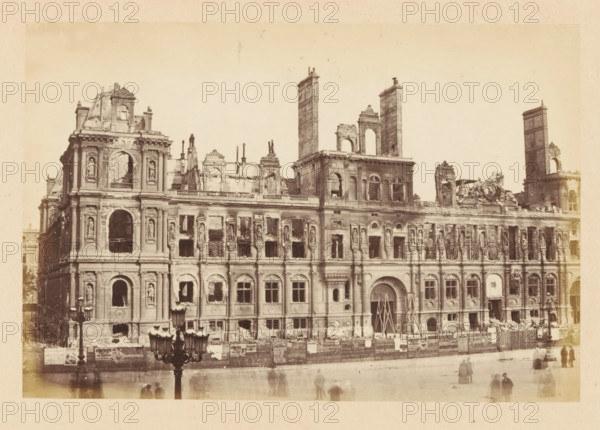 City Hall, View of the ruined Paris City Hall, photograph, page, 1871, photographer, Paris, paper, albumen print, height 101 mm x width 151 mm, ruin of a dwelling, house, castle, palace, A large stone building facade with ornate architectural details showing extensive damage and missing sections, multiple tall rectangular towers and partial walls rising above the roofline, rows of arched windows and niches with sculpted figures, broken openings and rubble along the base, scaffolding or temporary supports propped at a main arched entrance, a low fence or hoarding running along the front with posters attached, a streetlamp with multiple lanterns in the foreground, and several blurred figures on the pavement in front of the building.