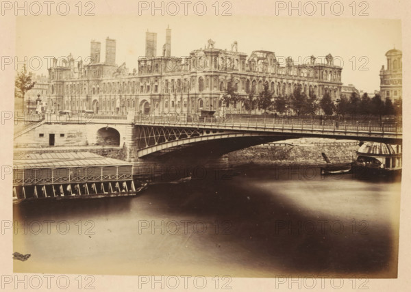 View of the destroyed Paris City Hall from the other side of the Seine, Hotel de ville, photograph, page, City Hall (Paris), 1871, photographer, Paris, paper, albumen print, height 101 mm x width 151 mm, river, ruin of a dwelling, house, castle, palace, A sepia-toned photograph shows a wide river in the foreground with smooth reflections, a stone embankment and an arched metal bridge crossing it, and an ornate multi-story classical building with numerous windows and sculptural details lining the riverbank behind the bridge; trees stand along the embankment partially obscuring parts of the building, a covered dock structure and a small boat with a domed shelter are visible at the water’s edge, and additional buildings and a solitary tree appear at the edges of the scene.