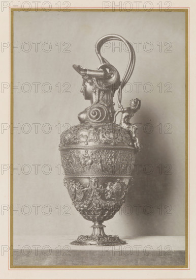 Lamppot of Emperor Charles V, seen from the side, Aiguière de Charles V (profile), Musée National du Louvre Galerie d'Apollon, page, photomechanical print, inscription: 'ateliers de reproductions artistiques', photographer: Léon Vidal, Paris, cliché maker: Léon Vidal, c. 1876 - c. 1883, Léon Vidal, photographer, cliché maker, paper, collotype, phototypie, height 260 mm x width 198 mm, container of precious metal, chasing, embossing (industrial process), A tall ornate metal vessel with a pedestal base sits centered on a flat surface against a plain backdrop, featuring a rounded body densely decorated with relief scenes and decorative motifs, a midsection band with emblematic shields and garlands, and an upper body with a molded lip. The handle rises high above the rim in a looping curve and attaches to a sculpted female head with braided hair and a helmet-like headdress that forms part of the spout area, while a small seated figure clings to the handle near the vessel's shoulder.