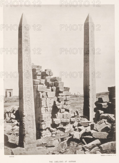 The obelisks at Karnak, page, photomechanical print, number: 'Plate XL', Karnak, cliché maker: Harroun & Bierstadt (possibly), New York (city), c. 1881, photographer, printer (person), North Africa, North America, paper, collotype, artotype, height 274 mm x width 205 mm, ruin of a building, architecture, obelisk, needle, temple, shrine, Egyptian religion, Two tall obelisks rise vertically from a rubble-strewn foreground, each with smooth tapering sides and pointed tops, their surfaces marked with vertical carved designs. Between and around the obelisks are piles of large, rough-hewn stone blocks stacked and scattered, creating irregular shapes and shadows. In the distant background a small rectangular stone structure with an open doorway stands on the plain, and a few thin palm trees are visible near the horizon. The sky above is pale and empty, filling most of the upper part of the image.