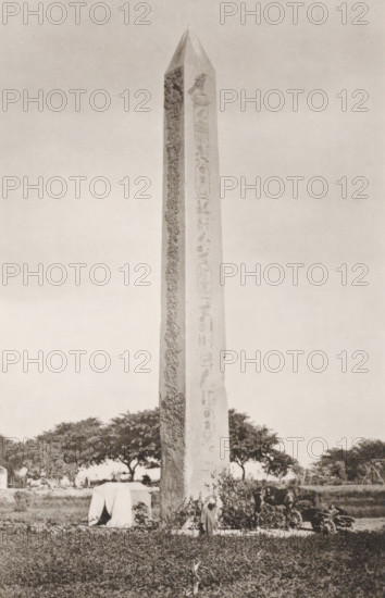 The obelisk at Heliopolis and Pompey's pillar at Alexandria, page, photomechanical print, number: 'Plate XLI', cliché maker: Harroun & Bierstadt (possibly), printer (person): Harroun & Bierstadt, New York (city), c. 1881, Harroun & Bierstadt, photographer, cliché maker, printer (person), North America, paper, collotype, artotype, height 270 mm x width 352 mm, Prints opposite page 124., column, pillar, architecture, obelisk, needle, North Africa, Alexandria, Pompey's Pillar, Serapeum, A tall stone obelisk occupies the right side of the image, showing a pointed top and faint carved markings along its shaft, while the left side shows a grassy field with scattered trees and a group of people and animals near the foreground, all beneath a pale sky.