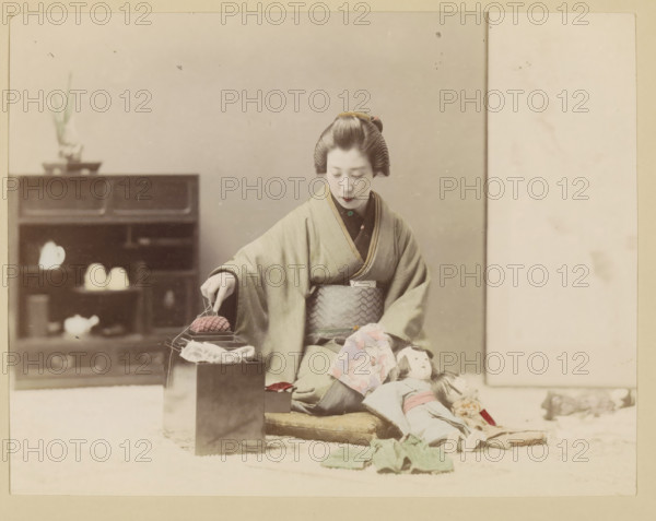 Woman making a doll, photograph, page, Japan, c. 1887 - in or before 1897, photographer, colorer (graphic design artist), paper, albumen print, hand coloring, height 186 mm x width 243 mm, Photo for page 57., adult woman, (playing with) dolls, interior, representation of a building, A seated woman in traditional dress with a patterned sash and styled hair looks downward while holding a small object above stacked boxes, alongside fabric pieces and a doll lying on cushions, with a low shelving unit and a small potted plant visible in the background.