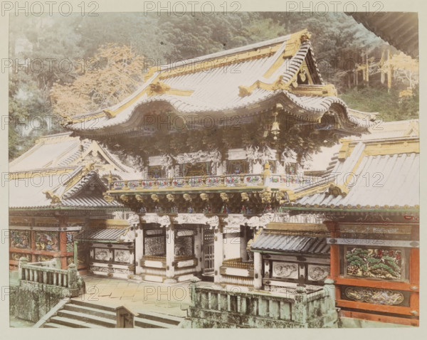 View of the Yomeimon Gate at Nikko, photograph, page, Nikko, Japan, c. 1887 - in or before 1897, photographer, colorer (graphic design artist), Kanto, paper, albumen print, hand coloring, height 186 mm x width 243 mm, Photo for page 63., facade (of house or building), gate, entrance, A large ornate traditional building with layered curved roofs topped with decorative ridge elements and gold-colored accents, intricate carved and painted wooden panels beneath the eaves, and sculpted brackets and ornamental details along the facade; stone steps and a weathered stone balustrade lead up to the entrance area, and painted panels with floral and nature motifs appear on the lower walls; trees and foliage form a background behind the structure.