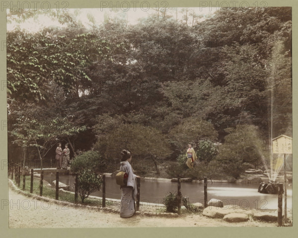 View of a pond in Osuwa Park in Nagasaki, photograph, page, Nagasaki, Japan, c. 1887 - in or before 1897, photographer, colorer (graphic design artist), Kyushu, paper, albumen print, hand coloring, height 188 mm x width 243 mm, Photo for page 135., public gardens, park, pond, pool, A landscaped scene with dense trees and shrubs in the background and a calm pond in the middle foreground, a narrow dirt path running along the front edge, wooden posts and rope forming a low fence beside the pond, a stone fountain sending a vertical stream of water into the pond near a cluster of rocks, a woman in traditional dress standing at the fence facing the water with a bundled item on her back, another woman in traditional dress standing on the far side of the pond among the bushes, and a pair of people standing together near a small bridge partially visible at one side.