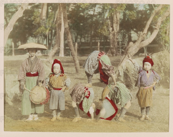 Child acrobats and their attendant in a park, photograph, page, Japan, c. 1887 - in or before 1897, photographer, colorer (graphic design artist), paper, albumen print, hand coloring, height 188 mm x width 243 mm, Photo for page 141., park, acrobat, circus, drum (musical instrument), Several children stand and bend on grass in front of trees and rocks, wearing striped robes and sashes in muted colors, some with red head coverings and one with a wide brim hat, one child holding a round drum with straps, others bent over or crouching with patterned cloths draped over them, and a child standing with a hand on a rock while another child stands with hands at their sides.