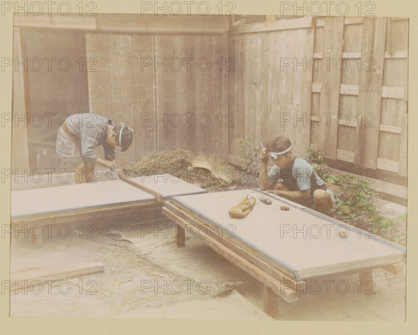 Two mat makers at work, photograph, page, Japan, c. 1887 - in or before 1897, photographer, colorer (graphic design artist), paper, albumen print, hand coloring, height 187 mm x width 243 mm, Photo for page 147., handicrafts, craftsman at work, door furniture, eg boot scraper, brush, doormat, A faded photograph shows two people working in a wooden courtyard with vertical plank walls. One person on the left is bent over a low platform, wearing a light-colored top and short skirt, with a cloth tied around their head. The other person on the right is seated on the ground near a corner of the enclosure, wearing a short-sleeved patterned top and a headband, holding something near their head. Several low wooden tables or boards are arranged across the foreground, one with a small bundle and a few small tools or objects on it. A pile of straw or dried grass rests against the wall between the two people, and some small plants grow near the seated person. The overall scene has muted, sepia-tone...