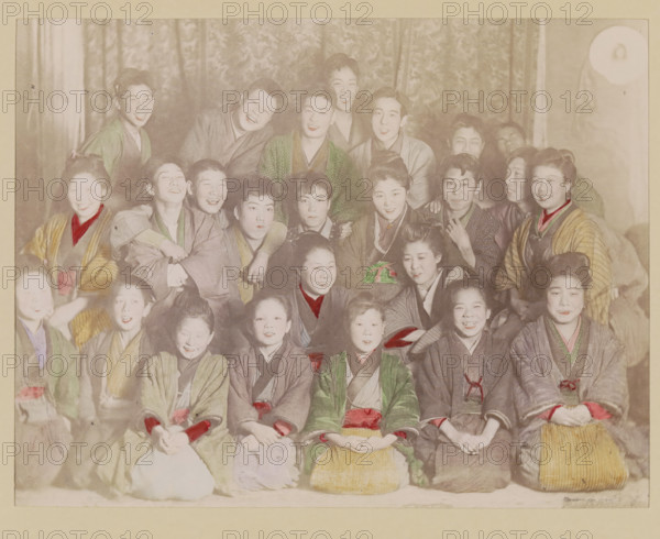 Group portrait of laughing boys and girls, photograph, page, Japan, c. 1887 - in or before 1897, photographer, colorer (graphic design artist), paper, albumen print, hand coloring, height 186 mm x width 242 mm, Photo for page 117., laughing, anonymous historical persons depicted in a group, in a group-portrait, A faded group portrait of many people arranged in rows against a patterned curtain backdrop, most wearing traditional robes with visible collars and sashes in muted colors, some robes showing hand-colored accents of red and green; the faces show a range of expressions from smiles to neutral looks, with hair styled in various updos and short cuts; hands rest on laps or shoulders, and a few people have arms around others; the image has an aged, sepia-toned appearance with slight discoloration and a pale border surrounding the scene.