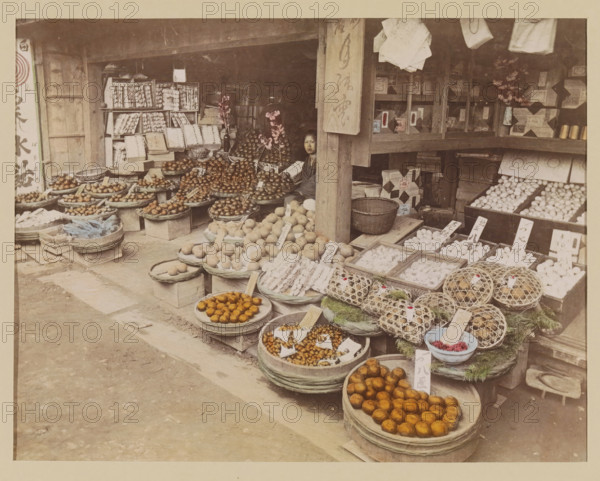 Display of foodstuffs at a shop, photograph, page, Japan, c. 1887 - in or before 1897, photographer, colorer (graphic design artist), paper, albumen print, hand coloring, height 187 mm x width 243 mm, Photo for page 123., shop, store, vegetables, A storefront with wooden beams and shelves is filled with round baskets and trays holding various fruits and goods, including piles of round fruits in different shades and textured items arranged on trays and in woven cages, with small rectangular markers placed among them; a seated person is partially visible behind a wooden post near the center, with a neutral facial expression; shelves and boxes line the back wall, some stacked and some displayed in open compartments, and a few vases with flowers sit on the shelves; the ground in front of the shop is a worn paved surface with scattered wooden boards and crates.