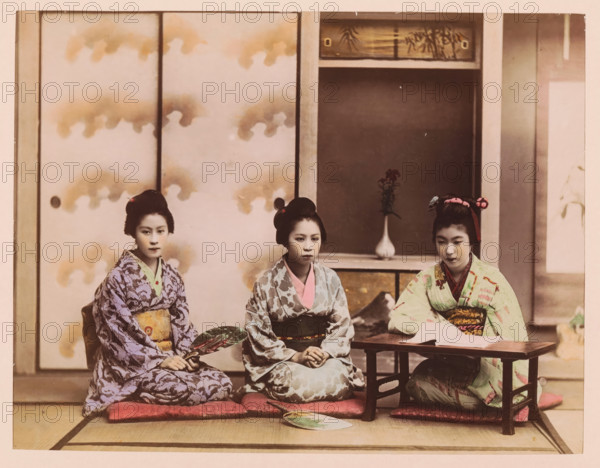 Three seated geishas in an interior, photograph, page, Japan, c. 1887 - in or before 1897, photographer, colorer (graphic design artist), paper, albumen print, hand coloring, photographie peinte, height 186 mm x width 242 mm, Photo for page 171., adolescent, young woman, maiden, fan, interior, representation of a building, Three young women wearing patterned kimonos sit on floor cushions in front of sliding panels, each with hair arranged in traditional styles and subtle hair ornaments; the woman on the left holds a decorative fan and wears a purple patterned kimono with a contrasting sash, the central woman wears a pale patterned kimono with a darker obi and has her hands folded in her lap, and the woman on the right wears a light kimono with colorful accents and sits at a low wooden table with papers on it; behind them a recessed alcove contains a vase with a single floral arrangement, and the background shows sliding doors with cloudlike designs and a wooden transom above.