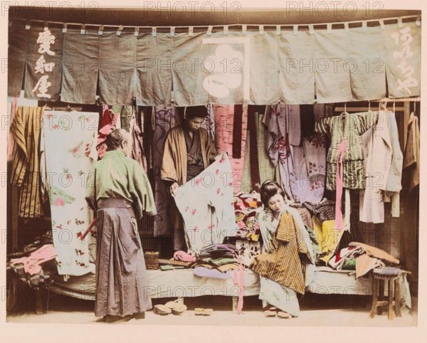 Display of merchandise in a second-hand clothing store, photograph, page, Japan, c. 1887 - in or before 1897, photographer, colorer (graphic design artist), paper, albumen print, hand coloring, photographie peinte, height 186 mm x width 242 mm, Photo for page 185., shop, store, textile fabric, cloth, fashion, clothing, A storefront scene showing fabric panels hanging from a horizontal beam across the top, with patterned and plain cloths draped and displayed behind and in front of the stall; three people are present, one standing with their back to the viewer wearing a long dark skirt and a light jacket, a second person standing inside the stall holding a pale patterned cloth and wearing a robe, and a seated person in a striped outer garment looking downward; piles of folded fabrics and garments rest on the bench in front of the stall and on the ground, with a small wooden stool and pairs of wooden sandals placed nearby; the overall color palette includes muted greens, pinks, yellows, a...
