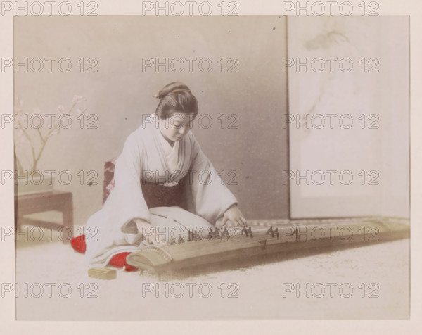Woman playing the koto, photograph, page, Japan, c. 1887 - in or before 1897, photographer, colorer (graphic design artist), paper, albumen print, hand coloring, photographie peinte, height 187 mm x width 243 mm, Photo for page 247., one person playing string instrument (plucked), interior, representation of a building, A seated woman wearing a traditional robe with long sleeves and a patterned sash is leaning forward with her hands on a stringed wooden instrument placed horizontally in front of her, her hair styled and pinned back, her gaze directed downward; a low wooden table with a potted flowering branch sits to her side, and a pale, softly textured backdrop with a vertical panel is visible behind her.