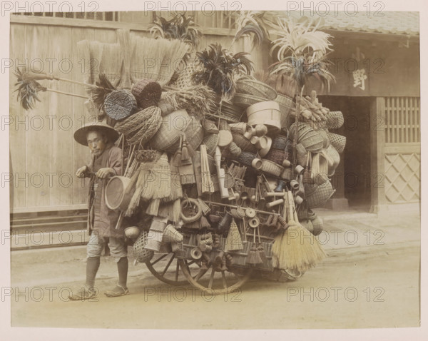 Itinerant merchant with a handcart and merchandise, photograph, page, Japan, c. 1887 - in or before 1897, photographer, colorer (graphic design artist), paper, albumen print, hand coloring, photographie peinte, height 186 mm x width 243 mm, Photo for page 253., merchant, salesman, hand-cart, A person wearing a wide-brimmed hat and traditional clothing stands beside a large wooden cart overflowing with woven baskets, brooms, straw hats, brushes, hand fans, cylindrical containers, and other woven and wooden household items, with several long-handled feather dusters and bundles protruding from the top; the cart has visible wooden wheels and is parked in front of a wooden building with slatted panels and a doorway partially obscured by a hanging curtain.