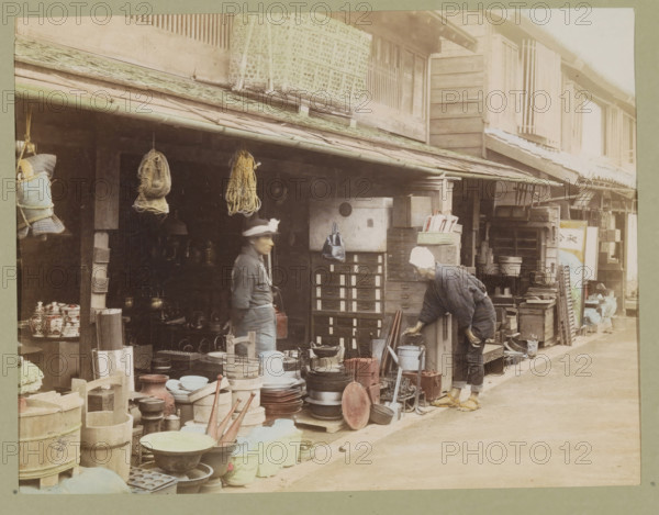 View of a shopping street, photograph, page, Japan, c. 1887 - in or before 1897, photographer, colorer (graphic design artist), paper, albumen print, hand coloring, photographie peinte, height 187 mm x width 243 mm, Photo for page 299., shop, store, street, shop-window, show-window, A street scene showing the front of a wooden shop with an overhanging tiled roof and various items displayed outside, including stacks of round and cylindrical containers, shallow bowls, trays, wooden buckets, metal pots, and other household wares arranged on the ground and on low platforms; two people stand near the shopfront, one facing sideways wearing a dark jacket and a hat with a brim, the other bending forward wearing a light head covering and dark clothing with sandals on their feet; hanging from the eaves are bundled ropes and wrapped objects, and a small bag is suspended near a column; the shop exterior features wooden shutters, small drawers and crates, and shelving filled with boxes and assorted...
