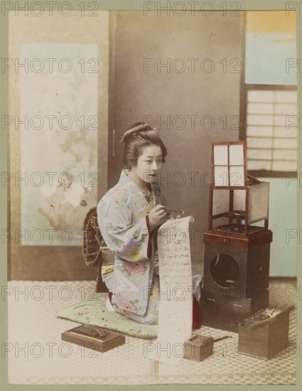 Woman writes a letter in an interior, photograph, page, photographer: attributed to Kusakabe Kimbei, Japan, c. 1887 - in or before 1897, colorer (graphic design artist), Attributed to, paper, albumen print, hand coloring, photographie peinte, height 243 mm x width 186 mm, Photo for page 281., letter, envelope, writing and letters, interior, representation of a building, A seated woman wearing a patterned kimono with floral designs and an ornate obi is holding a long scroll and a brush, her hair styled and adorned with hairpins; she sits on a cushion on a woven floor beside a small wooden box-like stand with a framed panel on top and other wooden containers and inkstone-like objects arranged nearby, with a painted screen and a window with grid panels visible in the background.