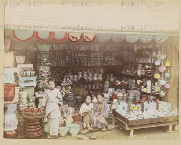 Shop with pottery, photograph, page, Japan, c. 1887 - in or before 1897, photographer, colorer (graphic design artist), paper, albumen print, hand coloring, photographie peinte, height 186 mm x width 243 mm, Photo for page 325., shop, store, container of ceramics: jar, jug, pot, vase, A storefront filled with stacked ceramics, bowls, jars, vases, plates, and teapots arranged on shelves, tables, and the floor, with patterned large pots and smaller cups in groups; hanging round decorative plates along the top edge and colorful hanging teapots on a rack to the right; a woman seated inside among the wares and three children in front of the display wearing patterned garments and sandals, one child standing while the others sit; baskets, trays, and stacks of dishes displayed on wooden platforms and crates, with various glazed finishes and floral designs visible.