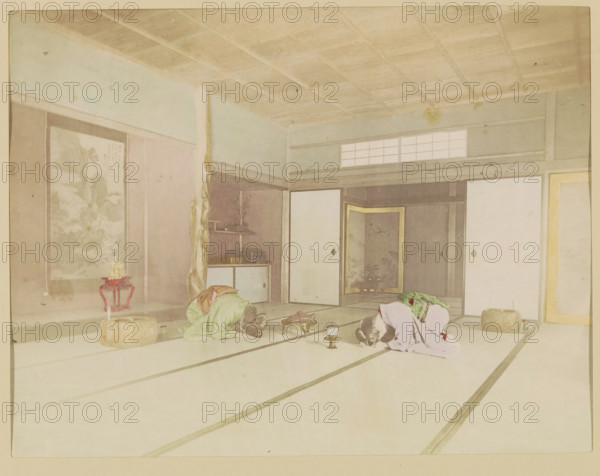Two women greet each other with a bow in an interior, photograph, page, Japan, c. 1887 - in or before 1897, photographer, colorer (graphic design artist), paper, albumen print, hand coloring, photographie peinte, height 187 mm x width 244 mm, Photo for page 315., kneeling figure, head bent forward, bowing - AA - female human figure, interior, representation of a building, An interior room with light-colored tatami flooring and wooden ceiling beams, sliding doors and panels along the walls, low furnishings including a small decorative stand with a floral arrangement and a round cushion, two women dressed in traditional robes kneeling and bowing with their heads lowered on the floor, a low tray with small vessels placed between them, a recessed alcove with decorative items and shelving, and soft, muted colors throughout the scene.