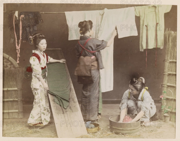 Three women washing kimonos, photograph, page, photographer: attributed to Kusakabe Kimbei, Japan, c. 1888 - in or before 1898, colorer (graphic design artist), Attributed to, paper, albumen print, hand coloring, photographie peinte, height 187 mm x width 244 mm, Photo for page 369., adult woman, dress, gown, wash house, home laundry, laundry room, Three women are engaged in household tasks in a muted indoor scene. One woman stands on the left wearing a patterned garment with a sash, her hair styled and adorned with a small hair ornament, and she rests a hand on a wooden incline draped with a striped cloth. A second woman stands center facing away, wearing a darker outfit with a tied sash and her hair in a bun, holding up a light-colored cloth that hangs from a horizontal pole. A third woman kneels on the right beside a wooden tub, sleeves rolled, working with a piece of cloth in the water, her hair tied back and a small floral detail near her. Hanging garments and ropes are visible on...