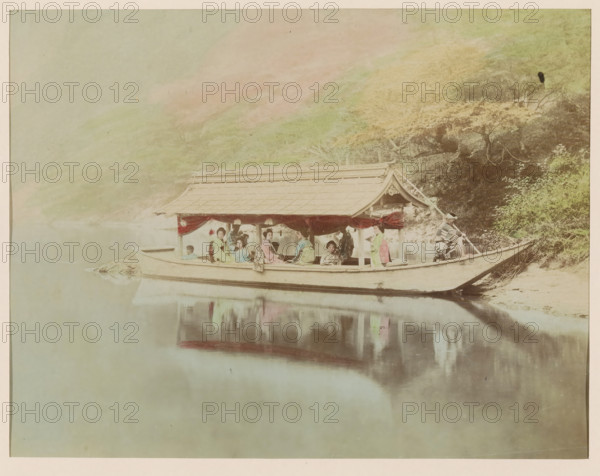 Houseboat, photograph, page, Japan, c. 1888 - in or before 1898, photographer, colorer (graphic design artist), paper, albumen print, hand coloring, photographie peinte, height 187 mm x width 244 mm, Photo for page 381., A long wooden boat with a roofed structure floats on calm water near a riverbank, with reflections visible on the water surface; several people sit inside the boat wearing traditional clothing in soft pastel colors, and red drapery hangs along the open sides of the roofed section; a person at the stern holds a pole and another person stands near the bow; behind the boat the bank shows trees and foliage with varied tones, and a gentle hillside rises in the background under a pale sky.