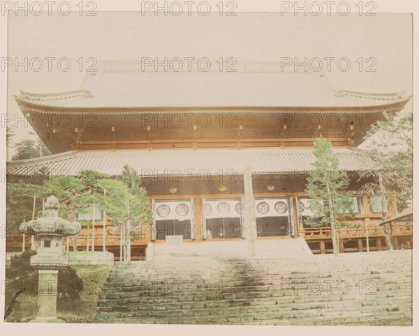 Front facade of the Manwangji Temple at Nikko, photograph, page, Nikko, Japan, c. 1888 - in or before 1898, photographer, colorer (graphic design artist), Kanto, paper, albumen print, hand coloring, photographie peinte, height 187 mm x width 242 mm, Photo for page 357., temple, shrine, Hinduism, Buddhism, Jainism, A large traditional wooden building with a tiered tiled roof fills the upper part of the image, showing ornate roof edges and eaves; beneath the roof a covered porch area with hanging circular decorations and wooden beams is visible, framed by wooden pillars and a central vertical post. Stone steps lead up toward the entrance in the foreground, flanked on the left by a weathered stone lantern and trimmed greenery, and on the right by several tall slender trees with green foliage. The scene has muted, slightly faded colors and a soft, even light across the structure and grounds.