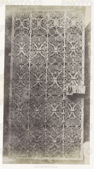 Porte en fer, Iron gate from the monastery of Le Puy-en-Velay, photograph, page, inscription: 'provenant du cloître de la Cathédrale du Puy.', number: 'PL. 13.', photographer: Hippolyte Malègue, France, c. 1850 - in or before 1857, paper, salted paper print, height 244 mm x width 135 mm, gate, entrance, ornament, circle and derived from circle, eg: guilloche, A vertically oriented image of an ornate metal gate filling most of the frame, showing a repeating pattern of spiral scrolls and floral-like motifs arranged in a grid, with vertical bars intersecting decorative elements and a rectangular metal latch attached on the right side; the gate is set within a lighter border and the overall image has a grainy, high-contrast texture.