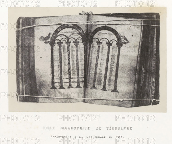 Bible manuscrite de Téodulphe, Bible from the cathedral of Le Puy-en-Velay, photograph, page, inscription: 'appartenant a la Cathédrale du Puy.', number: 'PL. 25.', photographer: Hippolyte Malègue, France, c. 1850 - in or before 1857, paper, salted paper print, height 136 mm x width 204 mm, Bible, An open book with visible pages showing decorative arches supported by columns, ornate capitals, and narrow vertical columns of small marks; small animal-like figures appear near the tops of the arches; the book edges and binding are visible along the sides, and light horizontal lines cross the image near the top and bottom margins.