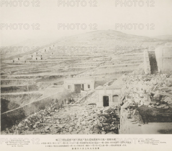 The rear defenses of the hill to the N. of the Tseszetai forts, on the W. Coast of Wei-Hai-Wei, page, photomechanical print, inscription: 'The road running by the telegraph post, to the left of the forts.', inscription: 'Expedition to Wei-hai-wei.', photographer: Ordnance Survey, Shandong, cliché maker: Kazumasa Ogawa, Japan, 1895-02-06, paper, collotype, photoprint, height 222 mm x width 286 mm, low hill country, fortress, defense, siege, A wide, pale landscape showing terraced hills with layered, horizontal ledges stretching across the scene, scattered with small rectangular openings in the slopes and rough rocky surfaces; in the foreground there are ruins and rubble leading to two stone buildings with arched doorways and flat roofs, one building partly buried and another framed by a taller stone wall and narrow steps, with a few indistinct human figures near the structures and a tall slender pole rising from the lower terraces.