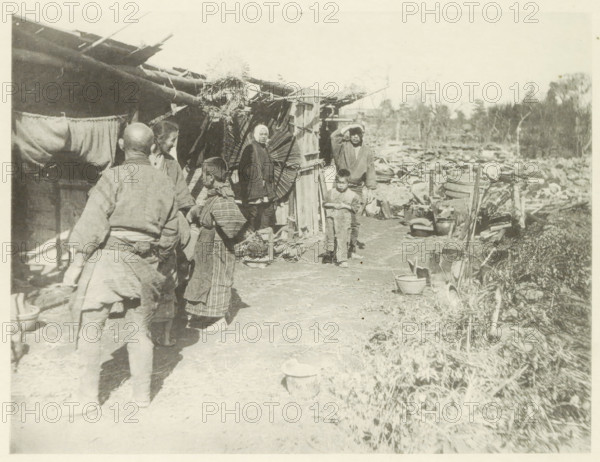 Temporary housing in Japan after an earthquake, page, photomechanical print, Japan, cliché maker: Kazumasa Ogawa, after 1891-10-28 - in or before 1892, photographer, paper, collotype, photoprint, height 198 mm x width 261 mm, housing, consequences of an earthquake, Mino–Owari earthquake 1891, A faded black-and-white scene showing several people standing and walking along a dirt path beside a row of makeshift wooden structures with thatched roofing and hanging cloths, with one person facing away from the camera in simple clothing and others grouped nearby in traditional garments, a child standing near the front looking forward, another person shielding their eyes with a hand, various household items and containers scattered along the path and near the structures, a large round woven object leaning against a wall, debris and more containers extending into the background toward a treeline under a pale sky.