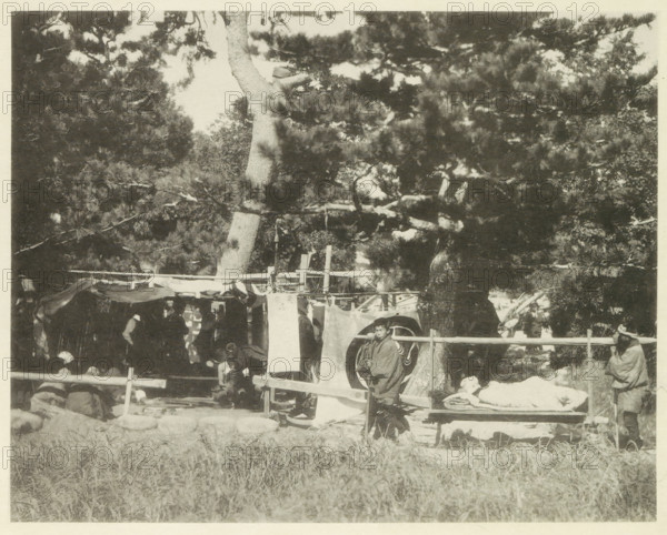 University field hospital in Japan after an earthquake, page, photomechanical print, Japan, cliché maker: Kazumasa Ogawa, after 1891-10-28 - in or before 1892, photographer, paper, collotype, photoprint, height 224 mm x width 283 mm, medical aid and nursing of the sick and wounded in a field-hospital, consequences of an earthquake, Mino–Owari earthquake 1891, A shaded outdoor scene shows large trees with dense branches and foliage filling the upper area and casting shadows below where a simple wooden structure with hanging cloth panels and draped fabric stands. Several people are visible around and within the structure, some seated and some standing, wearing layered garments and cloaks; one person stands near the front next to a circular patterned cloth, and another person sits or reclines on a low bench covered with folded textiles. A low wooden fence runs across the middle of the image, and grassy ground occupies the foreground. The overall image is monochrome with high contrast betw...