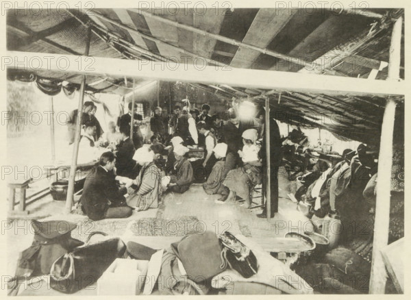 University field hospital in Japan after an earthquake, page, photomechanical print, photographer: Nakamura, Japan, cliché maker: Kazumasa Ogawa, after 1891-10-28 - in or before 1892, paper, collotype, photoprint, height 191 mm x width 264 mm, medical aid and nursing of the sick and wounded in a field-hospital, consequences of an earthquake, Mino–Owari earthquake 1891, A crowded interior scene under a sloped roof supported by poles, with a group of people seated and standing along a central aisle, many wearing head coverings and layered clothing, some seated on low stools or the floor and others standing and leaning; fabric, bags, and folded items are piled in the foreground, while benches, containers, and hanging items line the sides; the roof shows exposed beams and woven material, and light filters through openings creating bright patches and contrasts across the space.