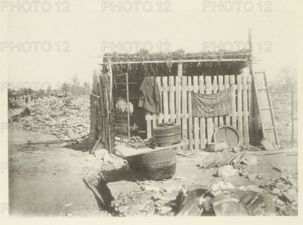 Temporary store in Japan after an earthquake, page, photomechanical print, Japan, cliché maker: Kazumasa Ogawa, after 1891-10-28 - in or before 1892, photographer, paper, collotype, photoprint, height 195 mm x width 275 mm, shop, store, ruin of a building, architecture, consequences of an earthquake, Mino–Owari Earthquake 1891, A weathered wooden shack with a thatched roof made of bundled vegetation stands at the edge of a debris-strewn dirt path, its front composed of vertical wooden slats and an open doorway revealing interior shelving and hanging rope; a coat hangs from a post and a patterned cloth is draped across part of the slatted wall, while various containers including stacked barrels, a round basin, baskets, and a board with a pile of round produce rest in front; a tall leaning ladder and scattered planks and rubble surround the structure, and a distant line of trees and indistinct figures can be seen along the horizon.