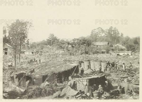 Destroyed buildings in Kasamatsu after an earthquake, page, photomechanical print, Kasamatsu, cliché maker: Kazumasa Ogawa, Japan, after 1891-10-28 - in or before 1892, photographer, Chubu, Gifu, paper, collotype, photoprint, height 211 mm x width 285 mm, ruin of a building, architecture, consequences of an earthquake, Mino–Owari earthquake 1891, A wide, desaturated scene shows an outdoor area cluttered with makeshift structures, thatch roofs, and scattered debris extending into a background of trees and low buildings; large cylindrical wooden tubs are grouped near the center right with small human figures near them, and more people are visible throughout the scene engaged in various activities; trees rise on the left and in the distance, and several simple houses and sheds are interspersed among vegetation; in the foreground there are bundles of straw, containers, baskets, and tools arranged near the huts, with poles and ropes stretching across parts of the image.
