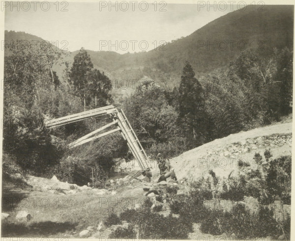 Destroyed bridge in the Neo Valley in Japan after an earthquake, page, photomechanical print, Japan, cliché maker: Kazumasa Ogawa, after 1891-10-28 - in or before 1892, photographer, paper, collotype, photoprint, height 227 mm x width 279 mm, bridge, dale, valley, consequences of an earthquake, Mino–Owari Earthquake 1891, A sepia-toned landscape showing a valley flanked by tree-covered hills and a distant ridge, with dense foliage of tall trees and shrubs in the midground and foreground. A wooden trestle structure leans diagonally over a small stream, with angled beams and rail-like planks visible. On a rocky slope near the stream a group of people are gathered, some standing and some seated, with packs and equipment on the ground nearby. The foreground shows grassy and rocky terrain with scattered stones and low vegetation. The sky above is pale and mostly clear.