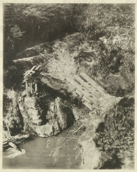 View of a destroyed bridge in the Neo Valley in Japan after an earthquake, page, photomechanical print, Japan, cliché maker: Kazumasa Ogawa, after 1891-10-28 - in or before 1892, photographer, paper, collotype, photoprint, height 283 mm x width 225 mm, bridge, dale, valley, consequences of an earthquake, Mino–Owari earthquake 1891, A monochrome photograph of a rocky riverside scene showing a wooden footbridge or planks laid across a steep bank, with stacked stones forming a retaining area beside the planks, rough rock outcrops, and a vertical wooden support structure perched on a ledge; water flows at the bottom with visible surface movement, fallen branches lie near the water, and vegetation including leafy trees and shrubs frames the scene with overhanging branches and textured foliage.