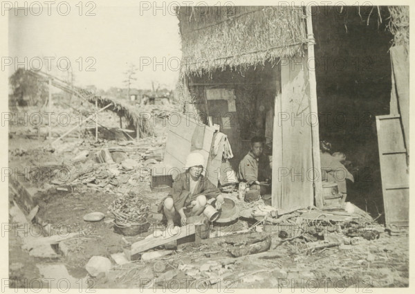 Temporary house in a village in Japan destroyed by an earthquake, page, photomechanical print, Japan, cliché maker: Kazumasa Ogawa, after 1891-10-28 - in or before 1892, photographer, paper, collotype, photoprint, height 196 mm x width 276 mm, ruin of a dwelling, house, castle, consequences of an earthquake, Mino–Owari Earthquake 1891, A faded photograph shows a thatched structure with a partially open doorway and a dark interior, while debris and scattered objects lie in front of it; a person wearing a light head covering and patterned clothing is crouched near a low wooden plank, holding a container, and another person is seated inside the doorway partially visible among household items and containers, with various baskets, bowls, planks, and bundles arranged on the ground around them.