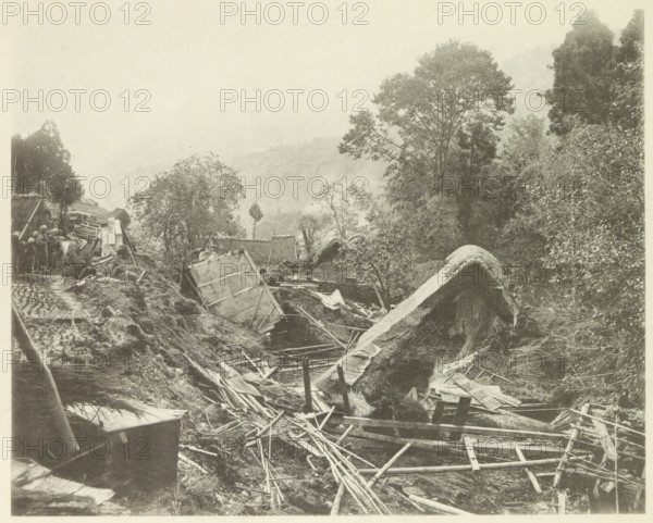 Destroyed houses in the Neo Valley in Japan after an earthquake, page, photomechanical print, Japan, cliché maker: Kazumasa Ogawa, after 1891-10-28 - in or before 1892, photographer, paper, collotype, photoprint, height 230 mm x width 288 mm, ruin of a building, architecture, consequences of an earthquake, Mino–Owari Earthquake 1891, A sepia-toned scene showing a collapsed rural area with broken wooden beams, planks, and ladders scattered across uneven ground, overturned thatched roofs and fragmented structures among mud and debris, dense trees and bushes rising on both sides with a large tree prominent on the right, a narrow muddy path or trench winding through the wreckage, a small group of people standing near upright remnants and stacked materials at the left edge, and distant hazy hills visible through the mist above the treeline.