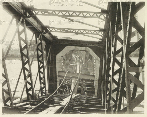 Collapsed railway bridge over the Nagara River in Japan after an earthquake, page, photomechanical print, Japan, cliché maker: Kazumasa Ogawa, after 1891-10-28 - in or before 1892, photographer, paper, collotype, photoprint, height 210 mm x width 266 mm, railway, tramway, rack railway, bridge, consequences of an earthquake, Mino–Owari earthquake 1891, A view through a metal truss bridge showing intersecting diagonal and horizontal beams forming repeating triangular patterns, riveted plates and girders, a central walkway with wooden planks and parallel rails, a man standing on the structure near the foreground facing away, layered arch-like openings framing the bridge interior, water visible beyond the bridge on either side, and distant vegetation along the far bank.