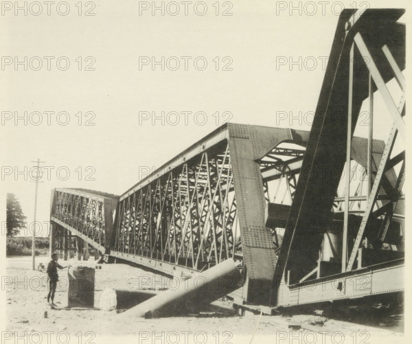 Collapsed railway bridge over the Nagara River in Japan after an earthquake, page, photomechanical print, Japan, cliché maker: Kazumasa Ogawa, after 1891-10-28 - in or before 1892, photographer, paper, collotype, photoprint, height 230 mm x width 278 mm, railway, tramway, rack railway, bridge, consequences of an earthquake, Mino–Owari earthquake 1891, A large steel truss bridge spans from the foreground into the distance with visible riveted girders and latticework, angled support beams and curved arch elements, and a section of heavy diagonal braces in the right foreground; a sandy ground area beneath the bridge shows scattered debris and a cylindrical concrete block with a person standing beside it holding a bicycle or similar two-wheeled object; a utility pole rises at the left edge and a small tree is visible near it, while the sky above is empty.
