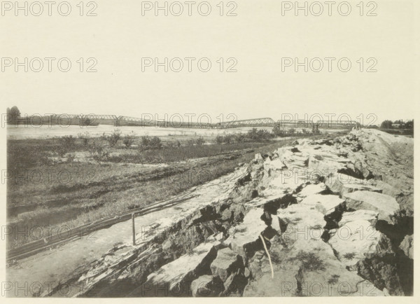 View of a collapsed railway bridge over the Nagara River in Japan after an earthquake, page, photomechanical print, Japan, cliché maker: Kazumasa Ogawa, after 1891-10-28 - in or before 1892, photographer, paper, collotype, photoprint, height 204 mm x width 282 mm, railway, tramway, rack railway, bridge, consequences of an earthquake, Mino–Owari earthquake 1891, A wide riverside scene with a long metal truss bridge spanning across the water in the distance, supported by piers and showing multiple connected spans; the near foreground shows a rocky embankment and uneven stone surface with scattered grasses and a narrow path or cleared strip alongside it; midground areas of low vegetation and patches of bare soil extend toward the riverbank; on the far shore there are trees and small clusters of foliage; the sky above is pale and mostly empty.