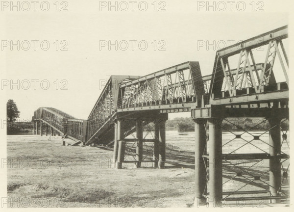 Collapsed railway bridge over the Nagara River in Japan after an earthquake, page, photomechanical print, Japan, cliché maker: Kazumasa Ogawa, after 1891-10-28 - in or before 1892, photographer, paper, collotype, photoprint, height 197 mm x width 287 mm, railway, tramway, rack railway, bridge, consequences of an earthquake, Mino–Owari earthquake 1891, A long metal truss bridge spans from the foreground to the background, showing repeating triangular framework and large vertical supports rising from the ground and water; the bridge sections slope and connect at different angles, with visible rivets and girders, and shadowed undersides above a mostly dry, textured riverbed; a solitary tree stands near the far bank, and distant foliage lines the horizon beneath a pale, empty sky.