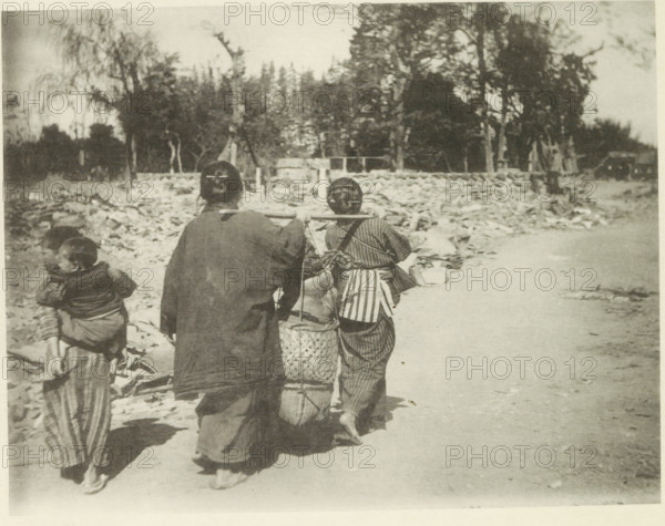 Two women carry belongings on a road in Japan after an earthquake, page, photomechanical print, Japan, cliché maker: Kazumasa Ogawa, after 1891-10-28 - in or before 1892, photographer, paper, collotype, photoprint, height 205 mm x width 265 mm, public road, carrying something with carrying-pole, consequences of an earthquake, Mino–Owari Earthquake 1891, Three people walk away from the camera along a dirt path beside a rubble-strewn area, two adults carrying woven baskets suspended from a wooden pole across their shoulders, one adult wearing a dark loose garment and the other wearing striped clothing with a sash; a child walks beside them with a smaller child carried on their back in a cloth, trees and scattered debris fill the background under a pale sky.