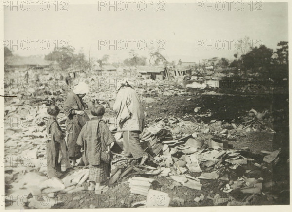 Two women with children stand among the destruction in Japan after an earthquake, page, photomechanical print, Japan, cliché maker: Kazumasa Ogawa, after 1891-10-28 - in or before 1892, photographer, paper, collotype, light print, height 181 mm x width 254 mm, ruin of a dwelling, house, castle, consequences of an earthquake, Mino–Owari Earthquake 1891, A sepia-toned photograph shows four people standing among a wide expanse of broken tiles and debris scattered across the ground, with three appearing to wear layered, loose garments and one wearing a head covering. The foreground is filled with irregular fragments of roofing tiles and curved ceramic pieces piled and strewn across dirt. In the midground, low wooden structures and scattered materials extend across the scene, with a few indistinct figures farther back. The background contains sparse trees, clusters of taller foliage, and more rubble extending toward a pale, empty sky.