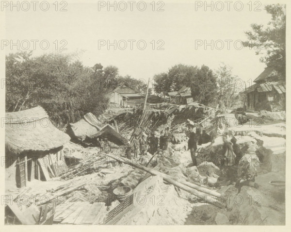Houses destroyed by an earthquake in Japan, page, photomechanical print, Japan, cliché maker: Kazumasa Ogawa, after 1891-10-28 - in or before 1892, photographer, paper, collotype, photoprint, height 221 mm x width 276 mm, ruin of a dwelling, house, castle, consequences of an earthquake, Mino–Owari Earthquake 1891, A sepia photograph shows a rural scene with uneven ground and scattered wooden beams and planks. Thatched roofs and simple wooden structures appear along the left and right edges, and a small building with a sloped roof sits amid trees in the middle distance. Trees and foliage occupy much of the upper left and right areas. Several people stand or sit among the debris and earth, some near a shallow trench and others on rocky ground. Woven baskets and round containers lie on the ground, and a leaning pole rises from the center of the scene. The overall view includes rough pathways and piles of earth and construction materials.