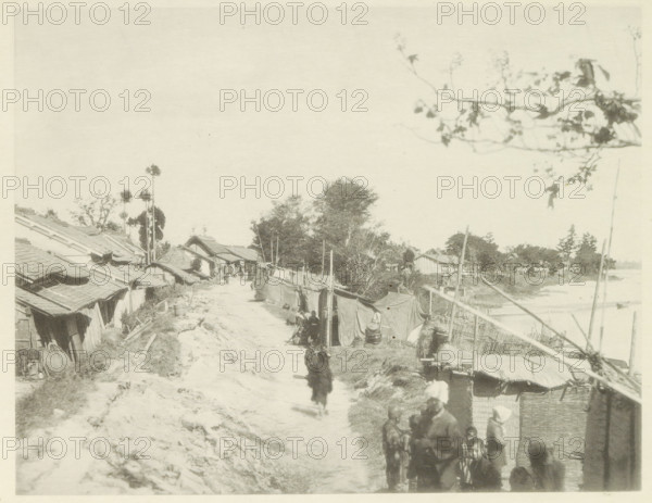 Temporary housing along the Biwajima River in Japan, page, photomechanical print, Japan, cliché maker: Kazumasa Ogawa, after 1891-10-28 - in or before 1892, photographer, paper, collotype, photoprint, height 223 mm x width 287 mm, ruin of a dwelling, house, castle, housing, consequences of an earthquake, Mino–Owari Earthquake 1891, A dirt path runs through a village lined with simple thatched and tiled roofs on one side and fabric-covered stalls and woven fences on the other, with several people walking and standing along the path and clustered near the foreground; trees and tall slender palms rise behind the houses toward the center, while a branch with sparse leaves frames the upper right, and a river or body of water is visible beyond the fences on the right.