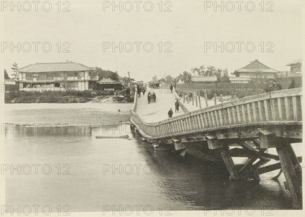 Collapsed bridge over the Biwajima River in Japan, page, photomechanical print, Japan, cliché maker: Kazumasa Ogawa, after 1891-10-28 - in or before 1892, photographer, paper, collotype, photoprint, height 196 mm x width 286 mm, bridge, consequences of an earthquake, Mino–Owari earthquake 1891, A wide wooden bridge extends from the foreground toward the distant shore, showing its slatted railing and supporting beams visible over water beneath, with several people walking along its length and a few standing near the far end; on the left bank there are low buildings with tiled roofs and rectangular windows set behind a grassy embankment, while the right bank features fenced yards, trees, and additional tiled-roof structures, and calm water fills the lower portion of the scene reflecting parts of the bridge.