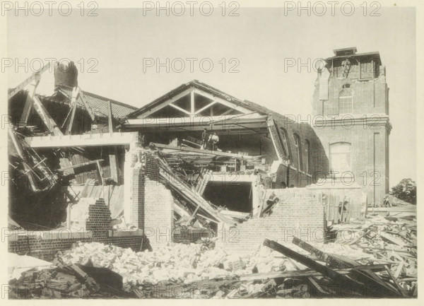 Collapsed cotton mill in Nagoya, page, photomechanical print, Nagoya, cliché maker: Kazumasa Ogawa, Japan, after 1891-10-28 - in or before 1892, photographer, Chubu, paper, collotype, photoprint, height 207 mm x width 290 mm, collapsing building, consequences of an earthquake, Mino–Owari earthquake 1891, A partially collapsed brick building with a gabled roof structure exposed, showing broken wooden beams, scattered bricks, and rubble in the foreground, two figures standing on a damaged interior floor amid debris, an intact tower section with arched windows and a damaged upper portion, leaning walls and exposed rafters, and piles of timber and masonry strewn across the site.